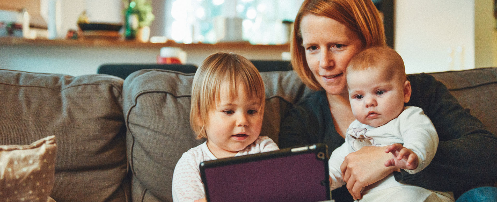 A woman sitting on the couch looks at a tablet with her two young children on her lap.